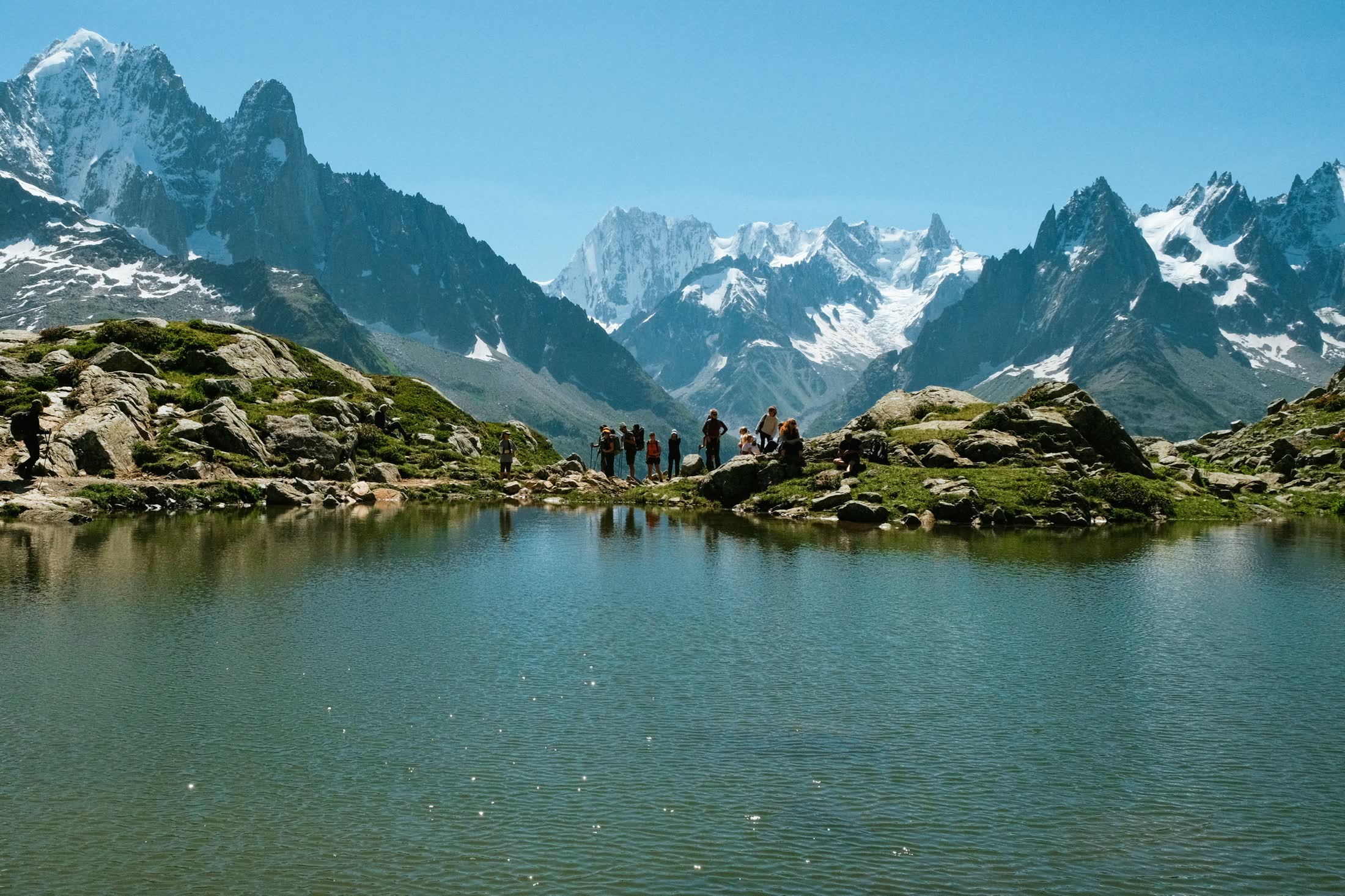 A group of hikers at an alpine lake beneath snowy peaks